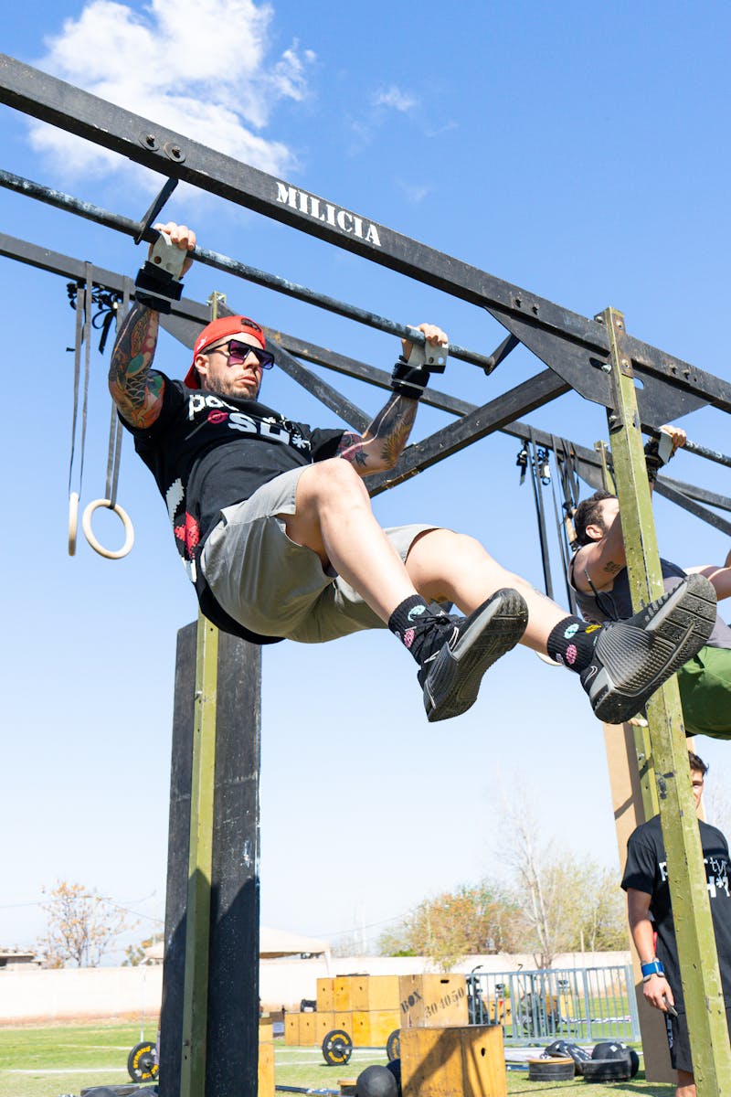 Athletes perform pull-ups on an outdoor obstacle course under blue skies.